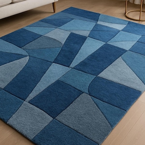 Könnte beinhalten: Rechteckiger Teppich mit geometrischem Muster in Blautönen. Das Design weist verschiedene Formen und Größen auf, wodurch ein moderner und strukturierter Look entsteht. Der Teppich liegt auf einem Holzboden, im Hintergrund ist ein Sofa zu sehen.