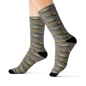 May include: Gray socks with a repeating pattern of yellow construction excavators and orange traffic cones. The socks have black heels and toes. The excavator design includes details like the cab, arm, and tracks.
