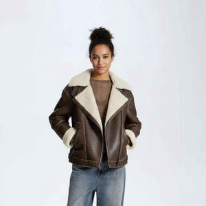 May include: A brown leather jacket with a cream shearling lining on the collar and cuffs. The jacket features an asymmetrical zip design and is worn with blue jeans. The model is wearing a sheer brown top.