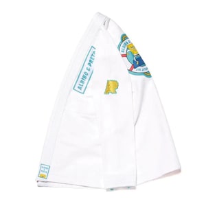 Könnte beinhalten: Weißer Jiu-Jitsu-Anzug mit dem Text "ALBINO & PRETA" in einer türkisfarbenen Box. Der Anzug hat einen gelb-grünen Aufnäher mit dem Buchstaben "R" und einen bunten Aufnäher mit einer Zeichentrickfigur. Der Anzug ist gefaltet.