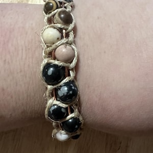 May include: A handmade bracelet featuring a series of natural stone beads in various colors, including black, brown, pink, and white. The beads are strung together with a natural fiber cord, creating a unique and earthy accessory. The bracelet is designed to fit a wrist.