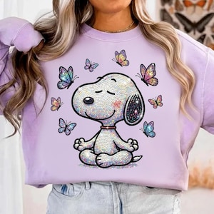 May include: A light purple sweatshirt featuring a cartoon Snoopy character in a meditative pose, surrounded by colorful butterflies. The Snoopy design is embellished with iridescent details, and the butterflies have vibrant, multi-colored wings. The sweatshirt is a casual, comfortable style.