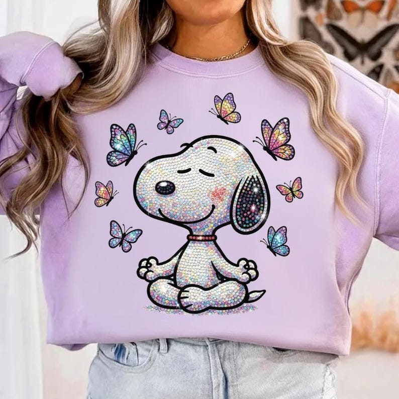 May include: A light purple sweatshirt featuring a cartoon Snoopy character in a meditative pose, surrounded by colorful butterflies. The Snoopy design is embellished with iridescent details, and the butterflies have vibrant, multi-colored wings. The sweatshirt is a casual, comfortable style.