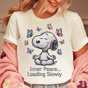 May include: White t-shirt featuring a cartoon Snoopy character in a meditative pose, surrounded by butterflies. The Snoopy design is embellished with iridescent details. The text "Inner Peace... Loading Slowly" is printed below the character.