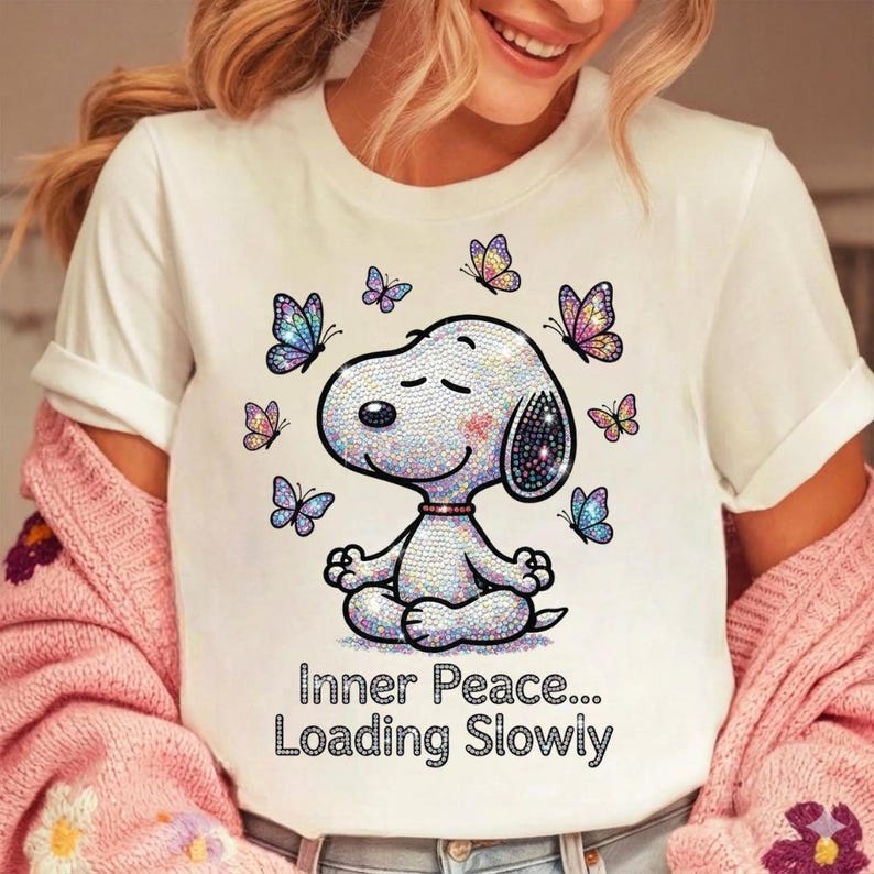 May include: White t-shirt featuring a cartoon Snoopy character in a meditative pose, surrounded by butterflies. The Snoopy design is embellished with iridescent details. The text "Inner Peace... Loading Slowly" is printed below the character.