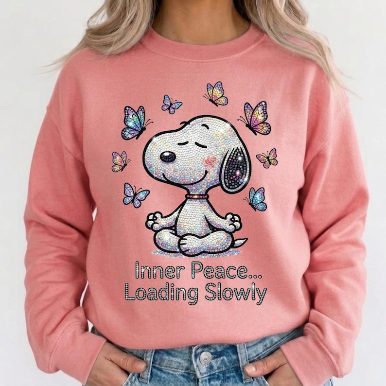 May include: A coral-colored sweatshirt featuring a rhinestone-studded Snoopy meditating with butterflies. The design includes the text "Inner Peace... Loading Slowly" in a sparkling font. The long-sleeved top is a casual style.
