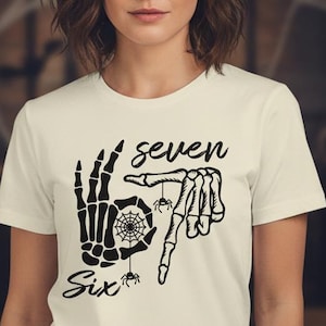 Pode incluir: Camiseta creme com um design de mão de esqueleto preto formando os números "7" e "6" com uma teia de aranha e aranhas. As palavras "Seven" e "Six" também estão impressas.