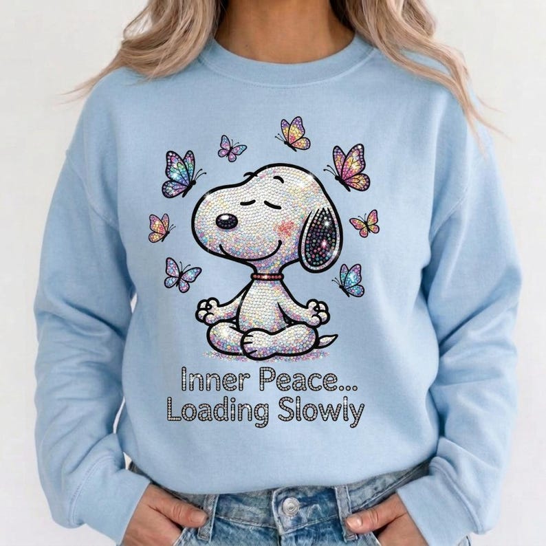 May include: Light blue sweatshirt featuring a sparkling Snoopy in a yoga pose, surrounded by colorful butterflies. The text "Inner Peace... Loading Slowly" is displayed below the character. A comfortable, casual top.