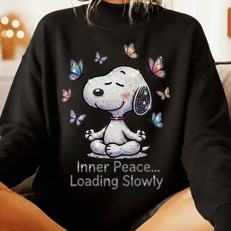 May include: Black sweatshirt featuring a sparkling Snoopy character in a meditative pose, surrounded by colorful butterflies. The text "Inner Peace... Loading Slowly" is displayed below the character. The design is embellished with iridescent rhinestones.