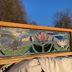 May include: Stained glass panel featuring a serene lotus flower design in shades of pink, white, and blue, set against a backdrop of green and iridescent glass. The artwork is framed in a natural wood border.