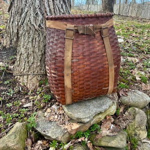 May include: A large, woven brown basket with a rectangular shape and a wide opening. It features two tan straps that go over the shoulders and a buckle closure. The basket is sitting on a pile of rocks near a tree.