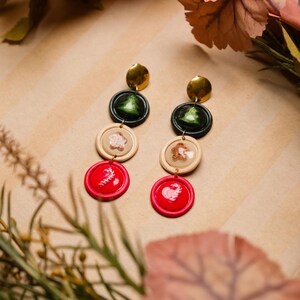May include: Dangling earrings with a festive theme. Each earring features a gold disc, a dark green circle with a Christmas tree, a beige circle with a snowflake, and a red circle with a candy cane. The earrings are designed for pierced ears.