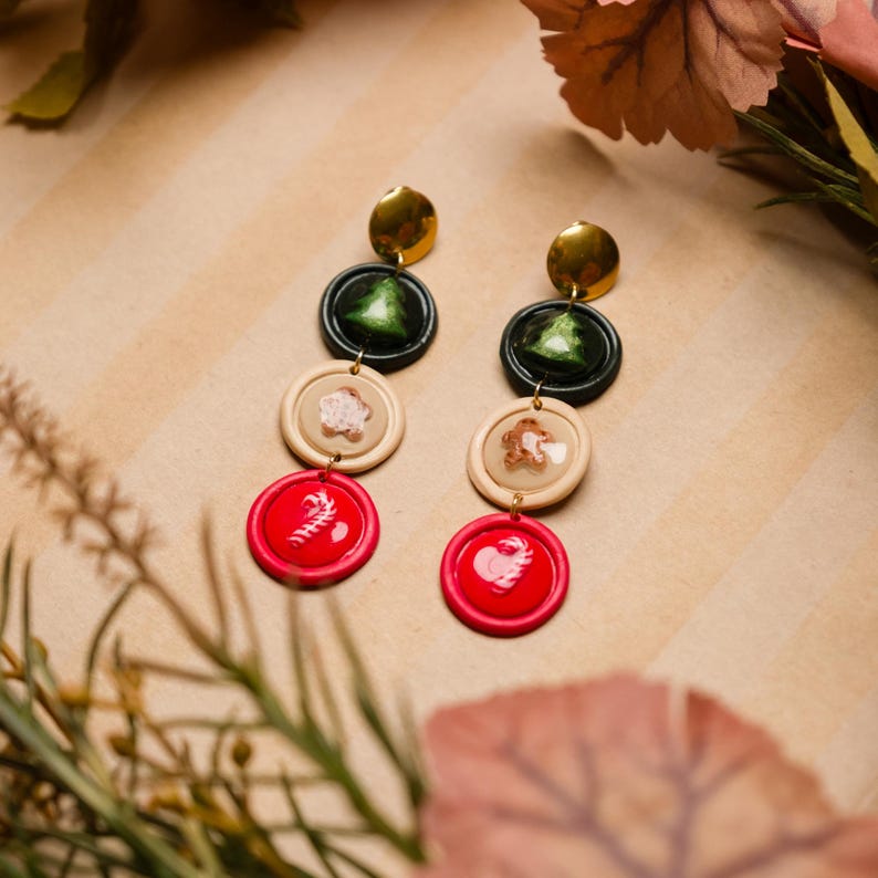 May include: Dangling earrings with a festive theme. Each earring features a gold disc, a dark green circle with a Christmas tree, a beige circle with a snowflake, and a red circle with a candy cane. The earrings are designed for pierced ears.