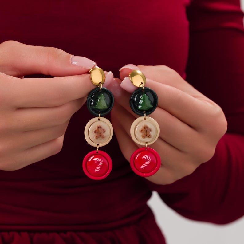 May include: Festive dangle earrings featuring a gold stud, a dark green circle with a green tree, a cream-colored circle with a gingerbread man, and a red circle with a candy cane design. These earrings are perfect for the holiday season.