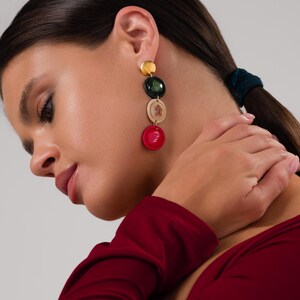 May include: Gold-toned dangle earrings with four colorful circular charms. The charms are in shades of gold, dark green, beige, and red. The earrings are worn by a person with dark hair and a red top.