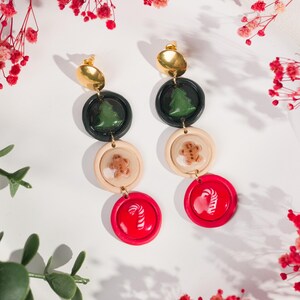 May include: A pair of festive earrings with gold-colored studs. Each earring features a black circle with a green Christmas tree, a beige circle with a gingerbread man, and a red circle with a candy cane design. The earrings are designed for the holiday season.