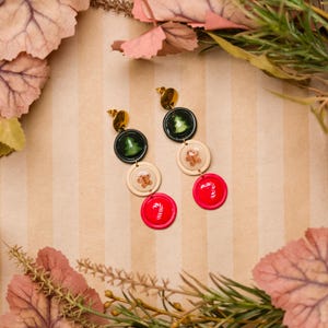 May include: Dangling earrings with a festive holiday theme. Each earring features a gold stud, followed by a dark green circle with a Christmas tree, a cream circle with a gingerbread man, and a red circle with a candy cane.