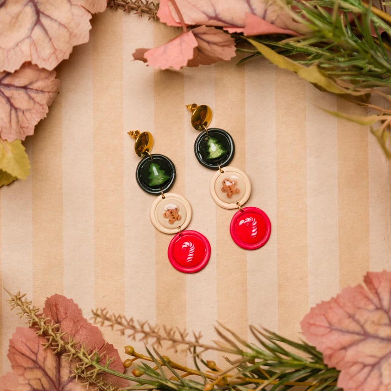 May include: Dangling earrings with a festive holiday theme. Each earring features a gold stud, followed by a dark green circle with a Christmas tree, a cream circle with a gingerbread man, and a red circle with a candy cane.