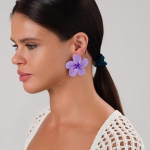 May include: A pair of large, lavender flower earrings. The earrings are shaped like five-petal flowers and are worn on the ear. The earrings are a vibrant shade of purple and are a statement piece of jewelry.