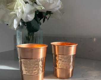 May include: Two cylindrical copper cups featuring a central hammered texture band bordered by beaded detailing. The cups have a smooth finish at the top and bottom rims, reflecting light with a warm metallic sheen, standing side by side.
