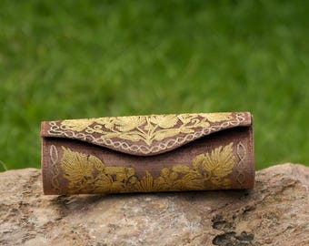 May include: A brown clutch with gold floral embroidery. The rectangular bag has a curved flap closure and is resting on a rock. The gold embroidery is detailed and covers the front and top of the clutch.