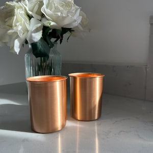 May include: Two cylindrical copper tumblers with a smooth, metallic finish and a straight-sided design. The cups have a warm, reflective orange-brown hue and a simple, unadorned rim, standing upright as a pair on a flat, light-coloured surface.