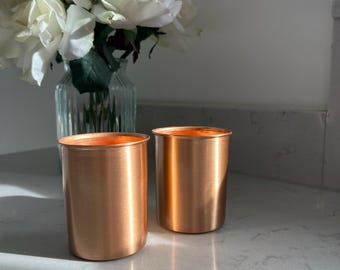 May include: Two cylindrical copper tumblers with a smooth, metallic finish and a straight-sided design. The cups have a warm, reflective orange-brown hue and a simple, unadorned rim, standing upright as a pair on a flat, light-colored surface.