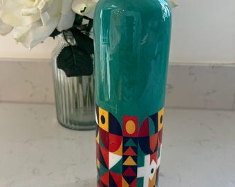 May include: A cylindrical water bottle with a metallic copper-colored cap. The upper portion is a solid teal color, while the lower section features a vibrant geometric pattern composed of red, yellow, blue, and white shapes on a dark background.