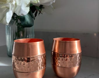 May include: Two matching copper tumblers with a rounded barrel shape. Each vessel features a central band with a hammered texture, bordered by rows of small raised dots. The metal has a polished, warm metallic finish that reflects light.