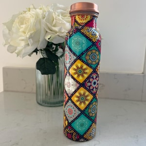 May include: A cylindrical water bottle featuring a vibrant, multi-coloured geometric pattern of diamonds filled with intricate mandala-style designs in shades of teal, yellow, pink, and white, topped with a metallic copper-coloured screw-top lid.