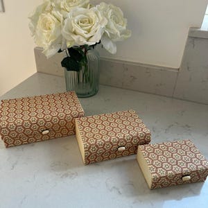 May include: Three rectangular boxes of varying sizes featuring a repeating geometric pattern in brown and cream. Each box has a small, light-coloured oval latch on the front. The boxes are arranged in a descending order of size from left to right.