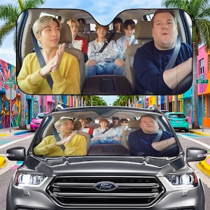 May include: Car sunshade featuring a group of people in a car, with a colorful street scene in the background. The sunshade shows a group of people in a car, with a man in a yellow sweater in the front seat. The car is a gray Ford.