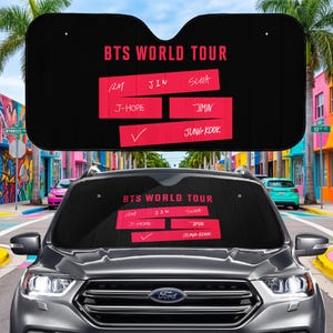 May include: Black car sunshade with the text "BTS WORLD TOUR" in red. The names of the band members are listed in red boxes. The sunshade is displayed on a car windshield in front of a colorful street scene.
