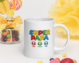 May include: White ceramic mug with the words "SUPER PAPA" in colorful block letters. Below the text are three mushroom graphics with the names "Lucas", "Raphael", and "Emma". The mug is surrounded by colorful candies and a yellow flower.