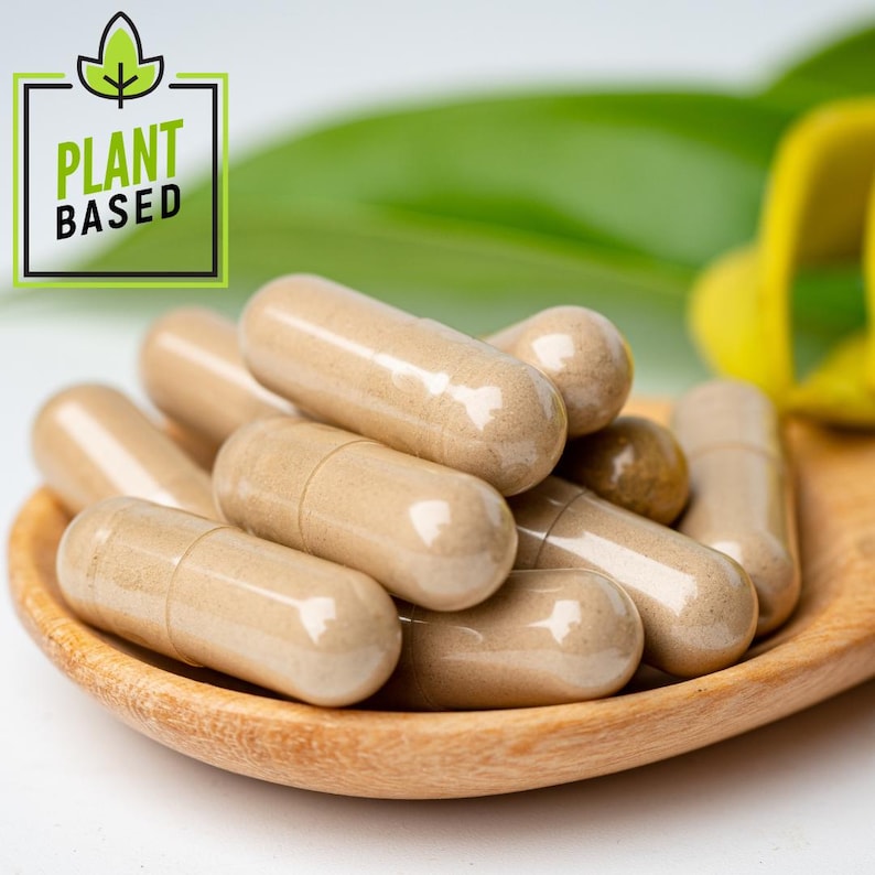 May include: Close-up of beige capsules in a wooden spoon. The capsules are stacked together, with a few visible. The spoon is set against a blurred background of green leaves and a yellow object. A green and black square with the words "PLANT BASED" is in the upper left corner.