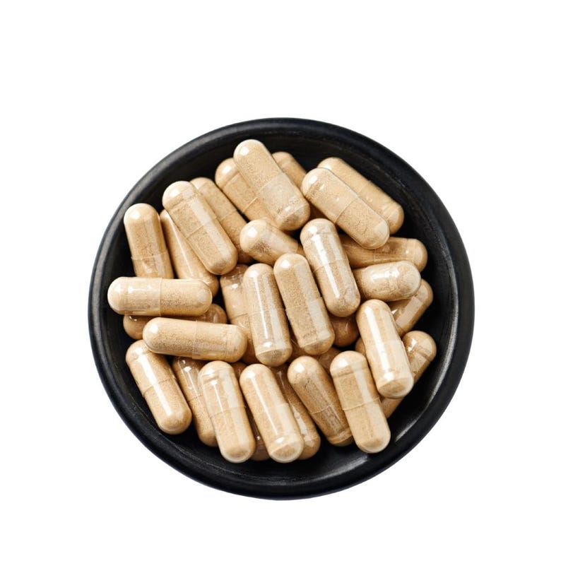 May include: A black bowl filled with numerous beige capsules. The capsules are cylindrical with rounded ends, and appear to be filled with a light-colored powder. The bowl is round and has a matte finish, contrasting with the bright white background.