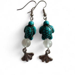 Puede incluir: Un par de pendientes colgantes con un dije de tortuga color turquesa con detalles en marrón oscuro, una cuenta redonda blanca esmerilada translúcida y un dije de hoja de ginkgo de metal plateado suspendido de un gancho de metal plateado.