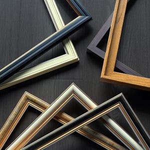 May include: An assortment of picture frames in various colors and styles. The frames include black, gold, cream, and wood tones. The frames are arranged on a dark wood surface, showcasing their different designs and finishes.