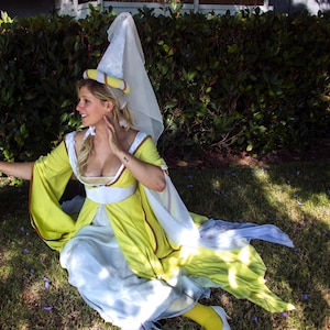 May include: A medieval-style costume in shades of yellow, white, and blue. The dress features long, flowing sleeves and a white veil. The person is sitting on the grass near a tree.