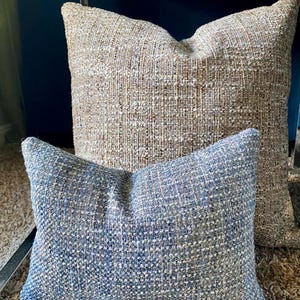 May include: Two decorative throw pillows. The larger pillow has a textured, woven fabric in shades of beige and brown. The smaller pillow is blue and gray with a similar woven texture. Both pillows are rectangular and appear plush.