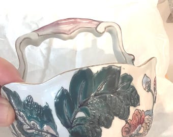 May include: White ceramic basket with a handle, featuring embossed green leaves and floral designs in pink and blue. The handle has a pink and white color scheme. A decorative piece for display.