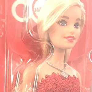 May include: A blonde doll with blue eyes and red lipstick, wearing a red dress with a silver necklace. The doll is packaged in a clear plastic container against a red background.