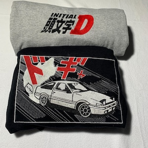 Puede incluir: Dos sudaderas dobladas. La superior es gris con "INITIAL 頭文字D" en negro y rojo. La inferior es negra con un gráfico de coche blanco y texto japonés en rojo.