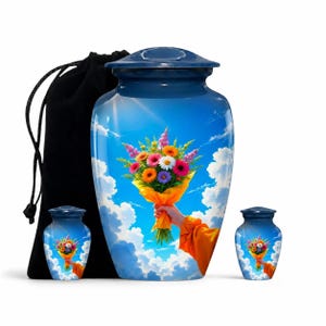 Op de afbeelding: Een grote blauwe urn en twee kleinere bijpassende aandenken-urnen, elk voorzien van een afbeelding van een hand die een kleurrijk boeket bloemen vasthoudt tegen een helderblauwe lucht met witte wolken. Een zwarte fluwelen zak is inbegrepen.
