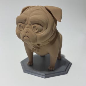 May include: A tan-colored pug dog figurine with a collar and bone-shaped tag, standing on a gray hexagonal base. The pug has large, expressive eyes and a slightly sad expression. The figurine is likely made of a 3D-printed material.