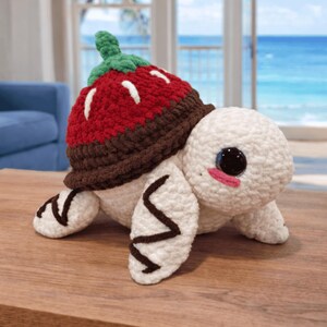 Peut inclure: Une tortue en peluche faite à la main, avec un corps blanc, de grands yeux noirs et des joues roses. La carapace de la tortue est une fraise rouge avec des détails blancs et une tige verte. Posée sur une surface en bois.