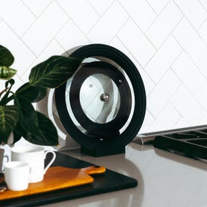 May include: A black and clear glass pot lid with a circular design. The lid has a black rim and a silver handle. The lid is on a gray countertop with a white tile backsplash. A wooden cutting board and white mugs are in the foreground.