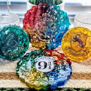 May include: A collection of colorful resin coasters and a small blue square, featuring various designs. The coasters display the Hogwarts crest, the Platform 9 3/4 logo, and house emblems like Slytherin and Hufflepuff. The items are arranged on a striped surface.