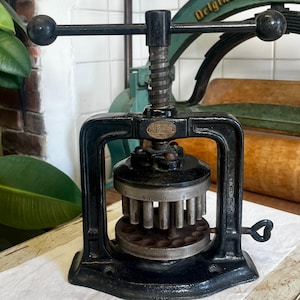 May include: Black antique metal press with a screw mechanism and a circular base. The press has a handle and a series of vertical rods. The word "Original" is visible on a green press in the background.