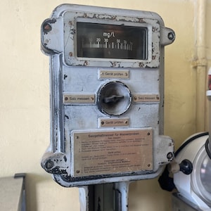 Puede incluir: Un medidor de sal industrial vintage, pintado en gris desgastado. Cuenta con un indicador con marcas en mg/L, un dial central y botones con texto en alemán. Una placa metálica proporciona instrucciones.
